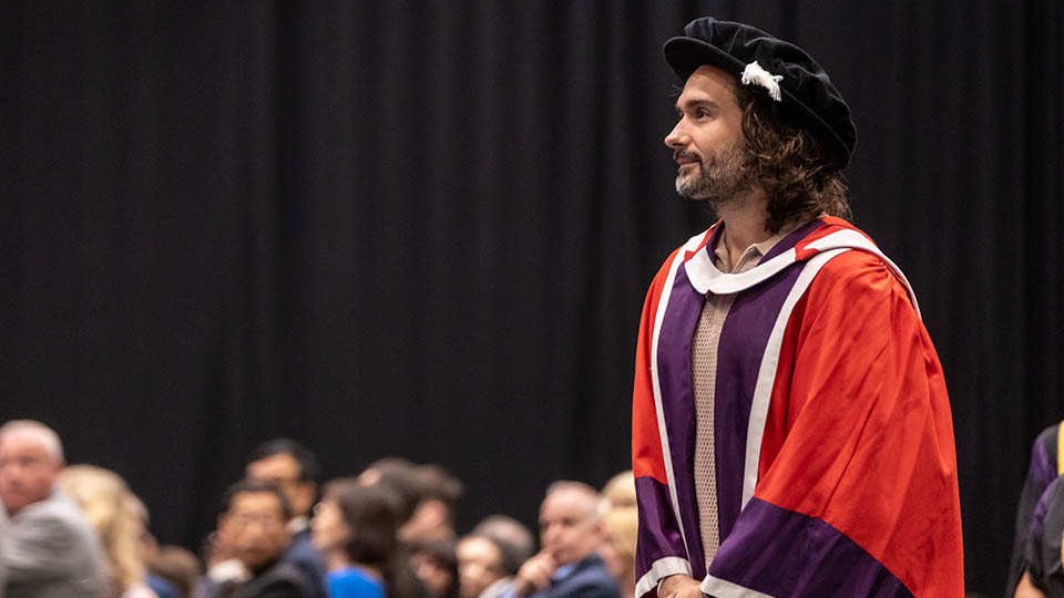 Joe Wicks wearing a red gown and black gap. There are people in the background as he walks past.