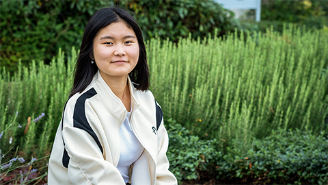 Student Tina is seated and there are plants behind her.