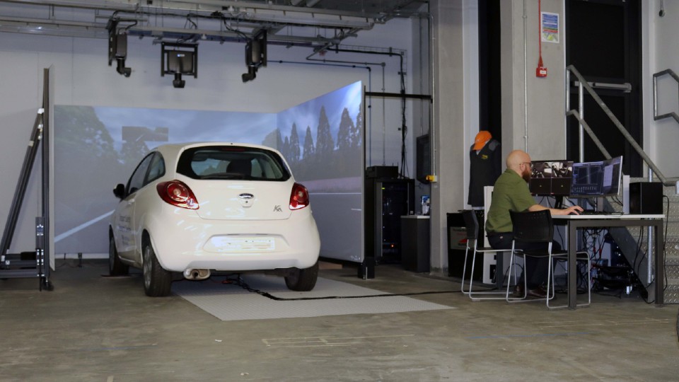 The driving simulator at Loughborough University, which comprises a white KA car surrounded by screens. Pictured on the screens is a road setting. There is a man sat to the right of the simulator operating a computer.