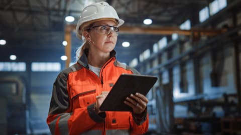 A worker in an industrial building, wearing a hard hat, hi vis clothing, and holding a tablet computer.