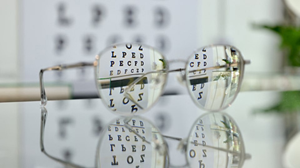 A picture of some spectacles with an eye-test reflected in the lens and visible on the wall behind.