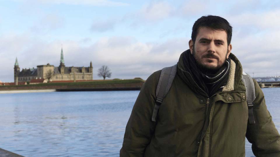 Student standing outside on a winter's day, smiling, with the sea and some buildings in the background