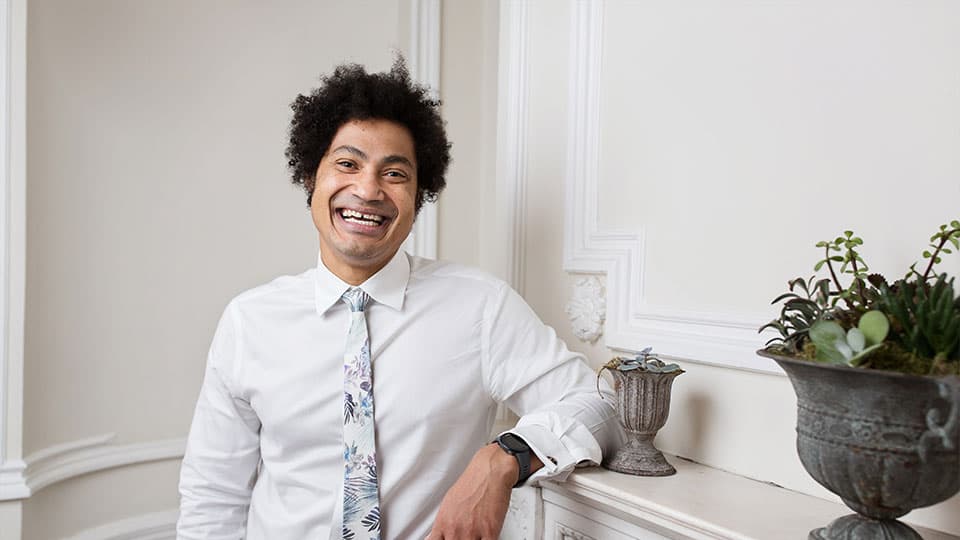 Jason is standing against a mantle piece smiling against a plain background with a plant pot in the bottom right hand corner.