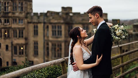 Joe and Sammy standing together looking into each others eyes on their wedding day and smiling. Warwick Castle is in the background blurred.