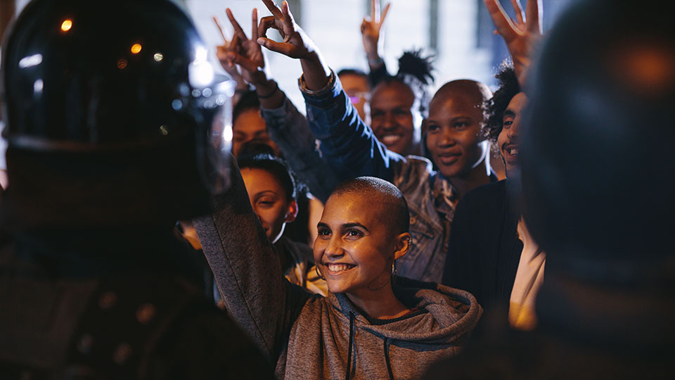 Photo of a woman in a crowd smiling with her arm up in the air and other people's arms in the air doing the victory sign with their hands,