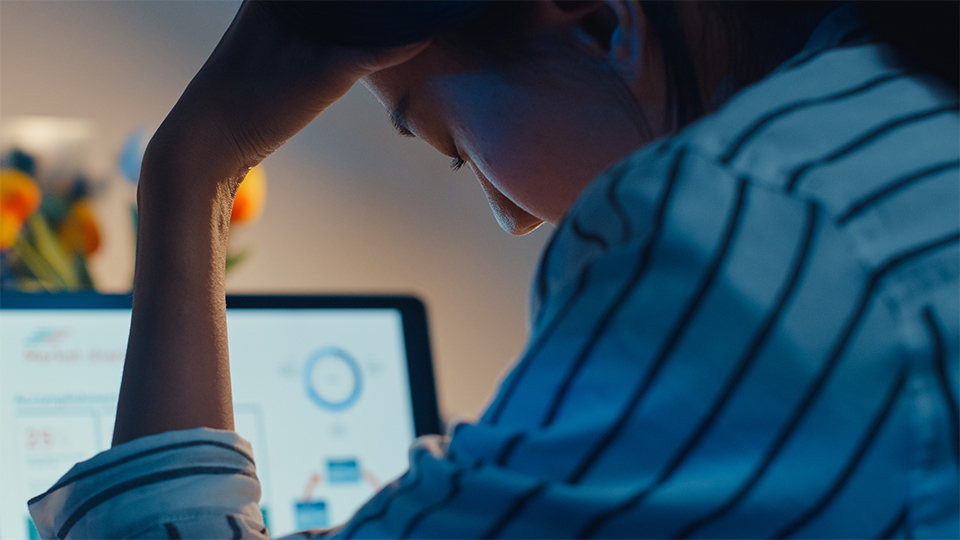 Close-up image of a woman with her head in her hand sat at a desk with a laptop switched on in the background