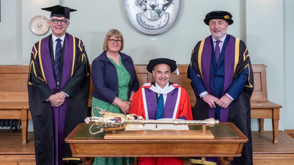 Three men wearing academic gowns and one woman wearing a dress and jacket. One man is sitting at a table with a ceremonial mace resting on top. 