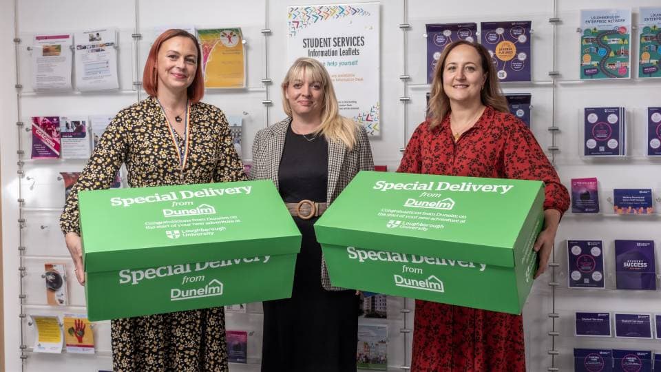 Three people standing in front of a wall of leaflets, two are holding big green boxes with Dunelm branding on.