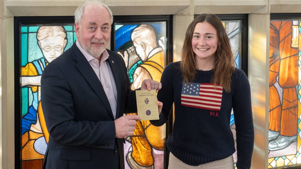 Zara being presented with a Gold Pin by the Vice-Chancellor Professor Nick Jennings. They are stood together smiling at the camera in front of stained glass windows