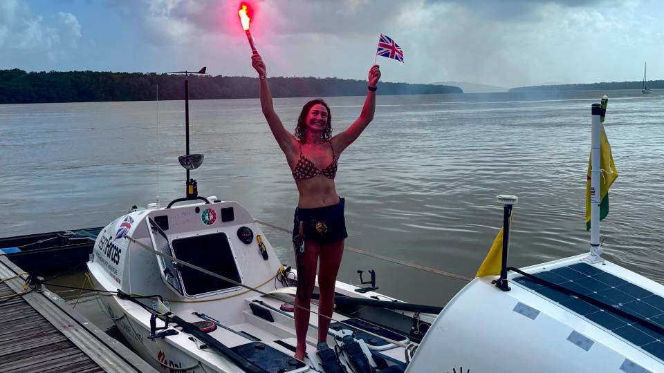 Zara standing on a rowing boat holding a red flare in one hand and a Union Jack flag in the other.