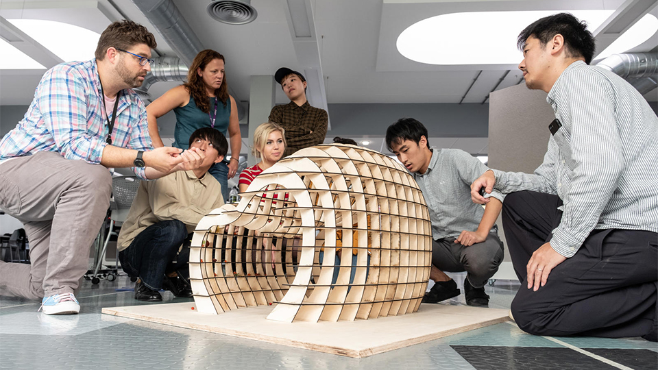 Photo of students from both universities and staff members looking at one of the models designed by a team at the Summer School