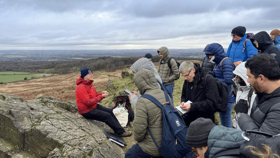 A group of people stood together at the Charnwood Forest Geopark listening to a man in a red coat sat down on a rock